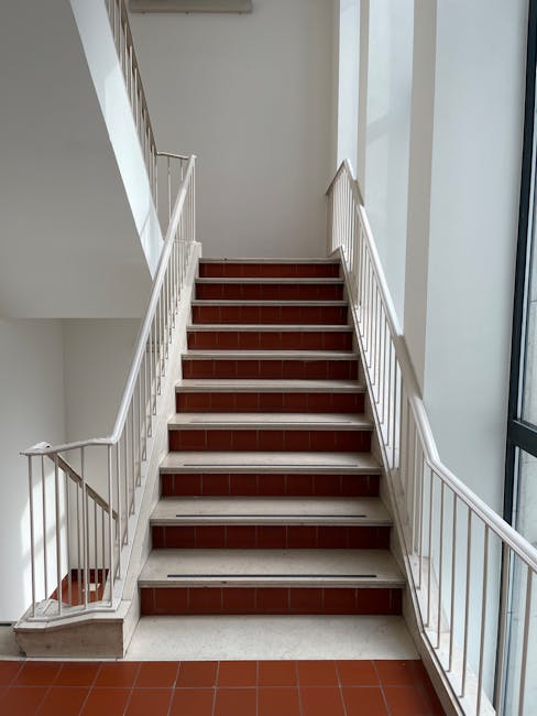 Indoor staircase with white metal railings and red-tiled steps, situated within a well-lit area adjacent to large windows allowing natural light. The steps are clean and free of debris, with a visible metal strip at the edge of each step for slip resistance. The surrounding walls are painted white, contributing to a bright and tidy appearance. The staircase appears to be in a residential or commercial building, possibly within a corridor or stairwell space. Holland Park Cleaner specializes in surface cleaning, deep cleaning, and sanitisation, ensuring high standards of hygiene for staircases like this one, maintaining cleanliness and safety within flats and communal areas.