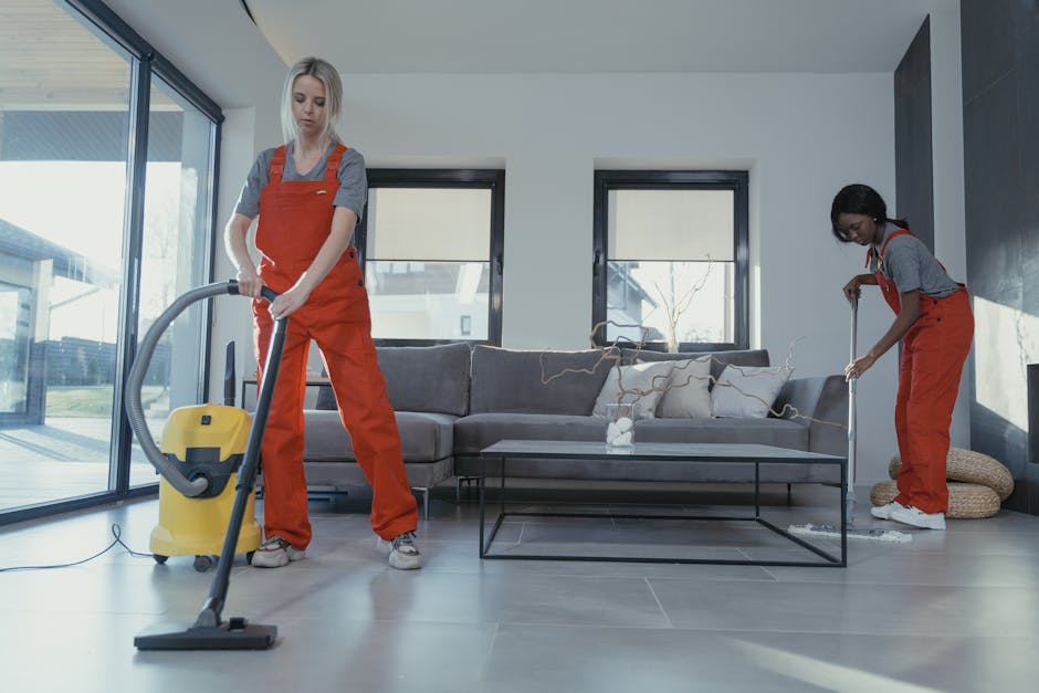 Two female cleaning professionals wearing orange uniforms and grey tops are performing surface cleaning in a modern living room. One is using a yellow vacuum cleaner on the tiled floor near large sliding glass doors, while the other is sweeping the sofa with a broom. The living room features a grey fabric sofa with cushions, a glass-topped coffee table, and large windows allowing natural light. The room's surfaces, including the tiled floor, sofa, and glass doors, appear clean and well-maintained, with minimal dust or clutter visible. The environment looks bright and organized, reflecting thorough domestic cleaning by Holland Park Cleaner.