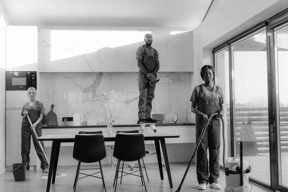 Two female cleaning professionals wearing orange uniforms and grey tops are performing surface cleaning in a modern living room. One is using a yellow vacuum cleaner on the tiled floor near large sliding glass doors, while the other is sweeping the sofa with a broom. The living room features a grey fabric sofa with cushions, a glass-topped coffee table, and large windows allowing natural light. The room's surfaces, including the tiled floor, sofa, and glass doors, appear clean and well-maintained, with minimal dust or clutter visible. The environment looks bright and organized, reflecting thorough domestic cleaning by Holland Park Cleaner.