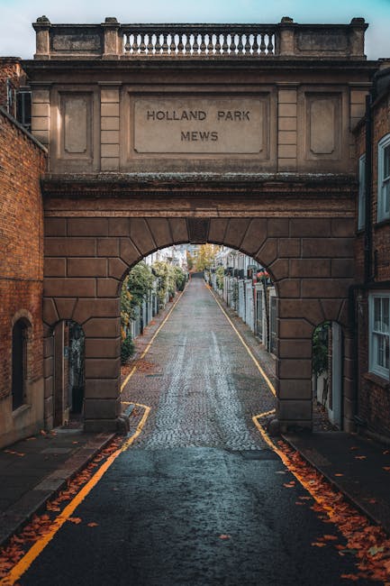 A cobblestone pedestrian pathway leading through a historic stone archway labeled 'Holland Park Mews,' flanked by residential buildings and garden fences. The pathway appears clean and well-maintained, with fallen autumn leaves along the sides. Bright natural light illuminates the scene, highlighting the texture of the stones and the greenery beyond. This image relates to surface cleaning and maintenance within residential areas, emphasizing the importance of cleanliness for hygienic and aesthetic purposes, as promoted by Holland Park Cleaner in their house cleaning guide for busy residents.