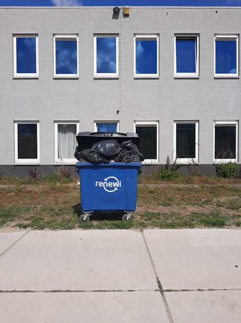 A street scene during daytime showing a worker wearing a traditional conical Asian hat, dark uniform, and reflective strips, pushing a large green waste bin with a foot pedal icon on the side. The wor