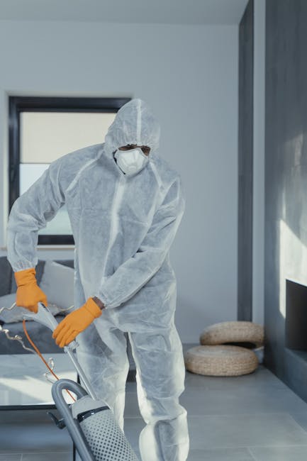 A professional cleaner dressed in a full white protective suit, face mask, and orange rubber gloves performs deep cleaning or surface sanitisation in a modern residential living room. The room features grey tiled flooring, a large window letting in natural light, and a minimalist grey wall. The cleaner is using a handheld device connected to a cord, possibly for spraying disinfectant, on the grey tiled floor. In the background, there are two round, beige cushions on the floor and a sleek, grey window frame. The scene emphasizes thorough domestic cleaning processes aimed at maintaining hygiene and cleanliness, consistent with services offered by Holland Park Cleaner in residential properties such as period homes in Holland Park.