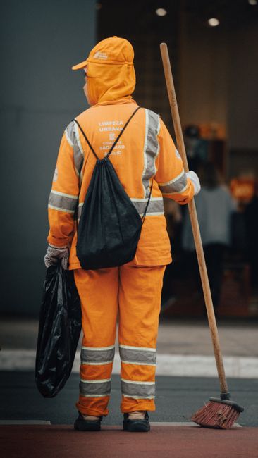 A commercial cleaner from Holland Park Cleaner is seen from above, wearing an orange high-visibility vest, grey protective overalls, red gloves, and black shoes, while pushing a green cleaning trolley
