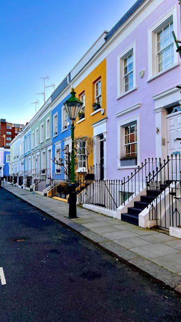 Photograph of a row of colorful Georgian-style terraced houses along a quiet street in Holland Park. The houses are painted in pastel shades including blue, yellow, and lavender, with white-framed sash windows and black wrought-iron railings and stairs. A classic black streetlamp is positioned in the foreground, illuminating the sidewalk. The scene is well-lit by natural daylight, highlighting the clean, well-maintained facades and the neatness of the street, which suggests a residential area serviced by professional cleaning services such as Holland Park Cleaner for thorough surface cleaning and maintenance of both exteriors and interiors.