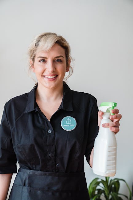 Two professional cleaning staff members from Holland Park Cleaner are standing back-to-back against a plain white wall, wearing matching beige uniforms with short sleeves. The woman on the left, with dark hair tied back, holds a vacuum cleaner hose in her right hand, while the woman on the right, with short blonde hair, is holding a mop handle with both hands, smiling at the camera. The scene suggests a focus on domestic cleaning or surface sanitisation, highlighting their readiness for deep cleaning tasks to ensure a hygienic environment in residential or commercial spaces. The clean, minimal background emphasizes their professional attire and cleaning tools, reflecting quality service available from Holland Park Cleaner.